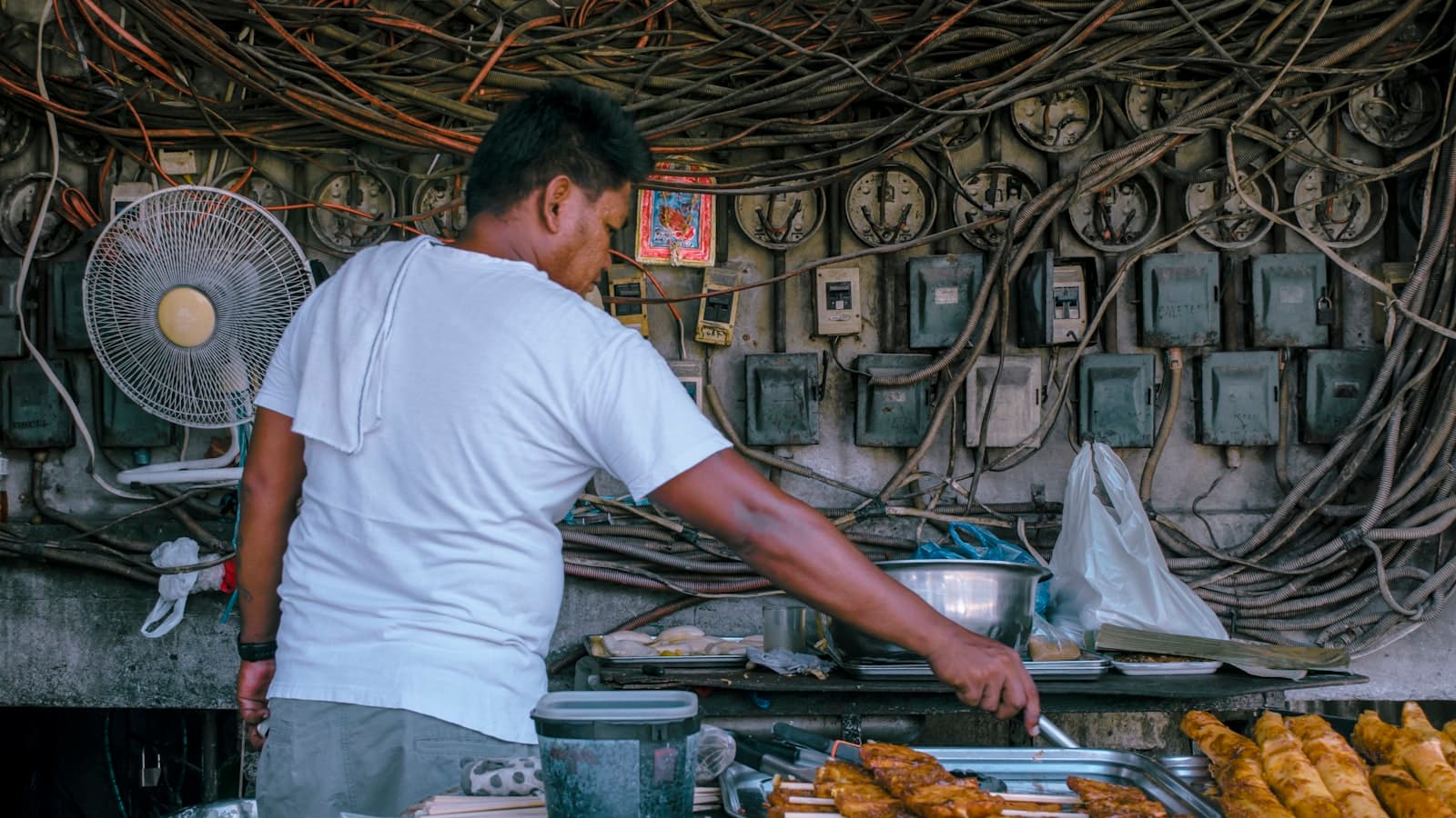 A Filipino street food vendor tending his grill stall in front of a dense tangle of electrical meters and wiring