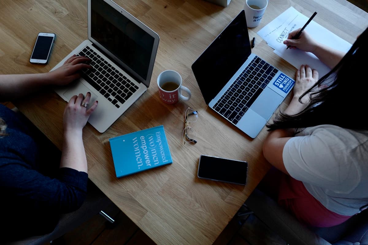 Two people working together on laptops at a desk, notes and a coffee mug between them